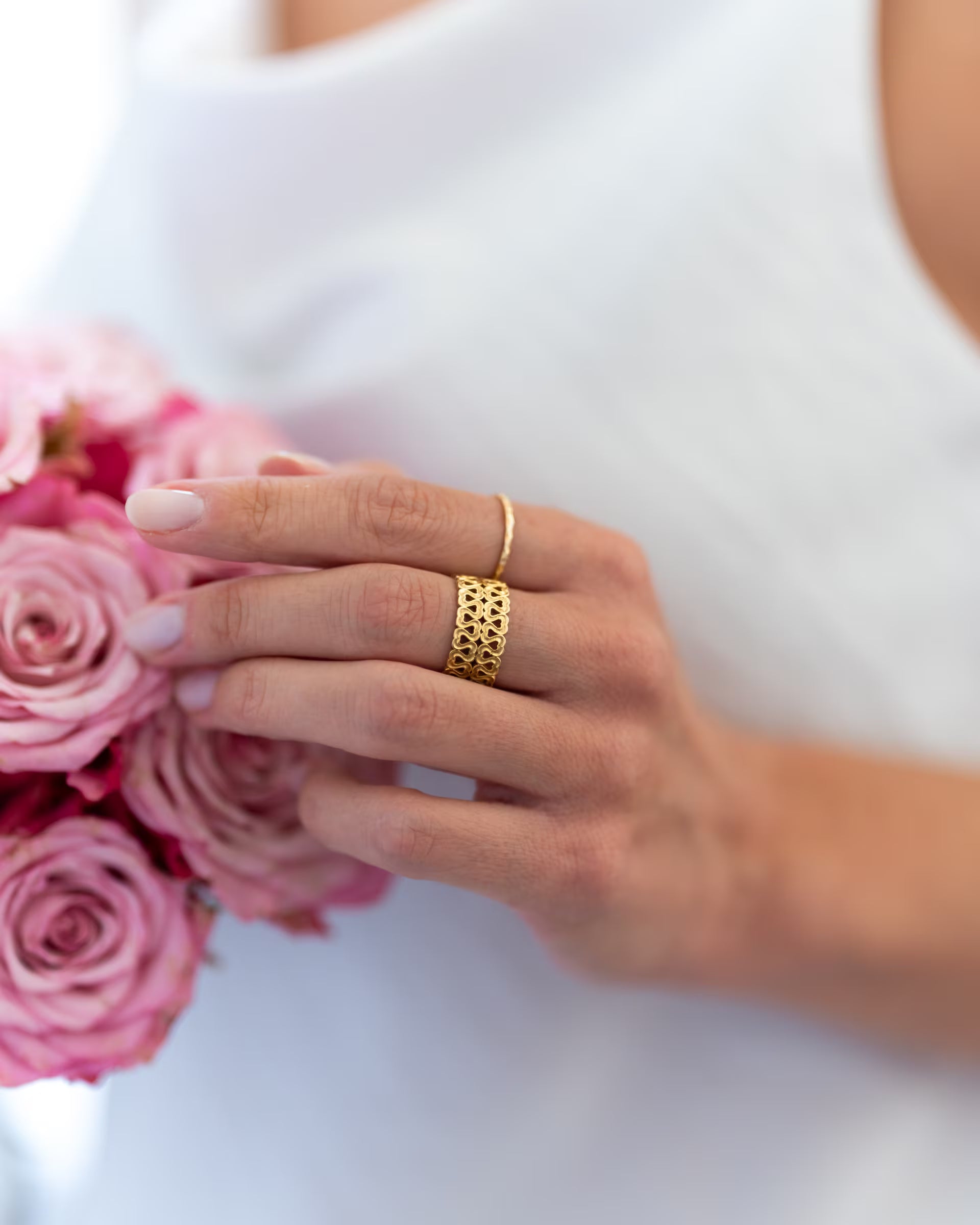 Romantische Nahaufnahme im eleganten Bridal-Look von einer Frau im weißen Kleid, deren Hand sanft an einem Strauß aus rosa Rosen ruht und eine Kombination aus auf Bali handgefertigtem goldenem Schmuck – bestehend aus dem breiten, offenen ENDLESS LOVE RING mit filigranem Herz-Muster sowie dem feinen, gehämmerten SARI STACKING RING – aus 18k vergoldetem 925er Silber von der Schmuckmarke ILUH trägt.
