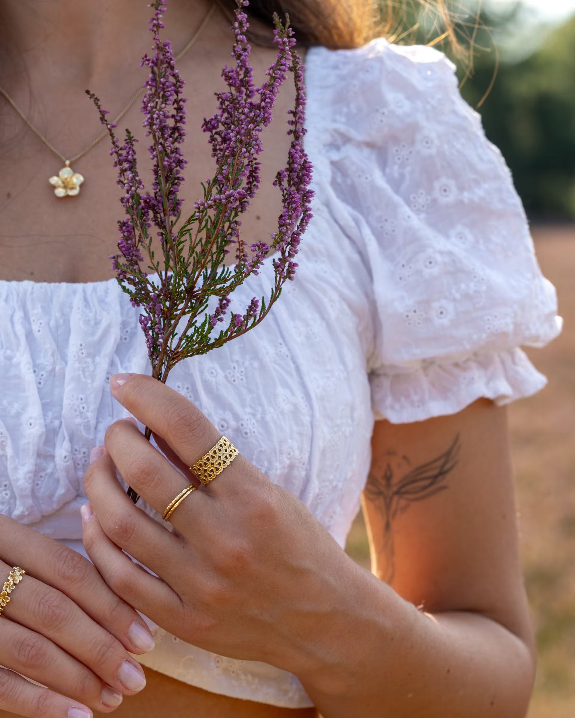 Nahaufnahme im romantischen Stil von einer jungen Frau im weißen Spitzenoberteil, die einen lila Heidekraut-Zweig hält und eine Kombination aus auf Bali handgefertigtem goldenem Schmuck – darunter die FRANGIPANI KETTE mit Blüten-Anhänger, den ENDLESS LOVE RING mit Herzmotiven und die feinen SARI STACKING RINGE – aus 18k vergoldetem 925er Silber von der Schmuckmarke ILUH trägt.