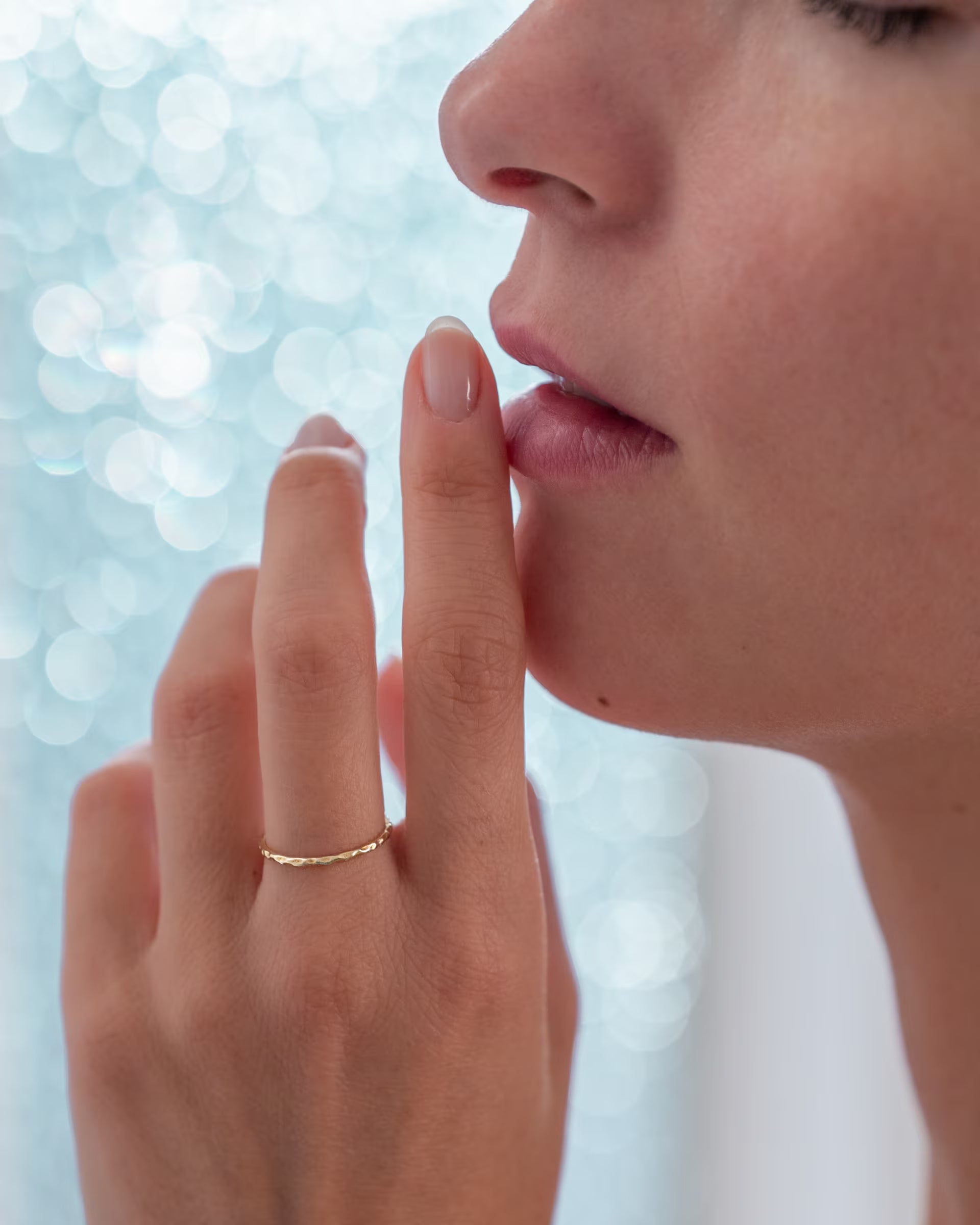Sinnliche Nahaufnahme im eleganten Stil von einer Frau im Profil, die nachdenklich einen Finger an die Lippen legt und den auf Bali handgefertigten goldenen, gehämmerten SARI STACKING RING aus feinem, 18k vergoldetem 925er Silber von der Schmuckmarke ILUH am Ringfinger trägt, vor einem sanft glitzernden, hellblauen Bokeh-Hintergrund.