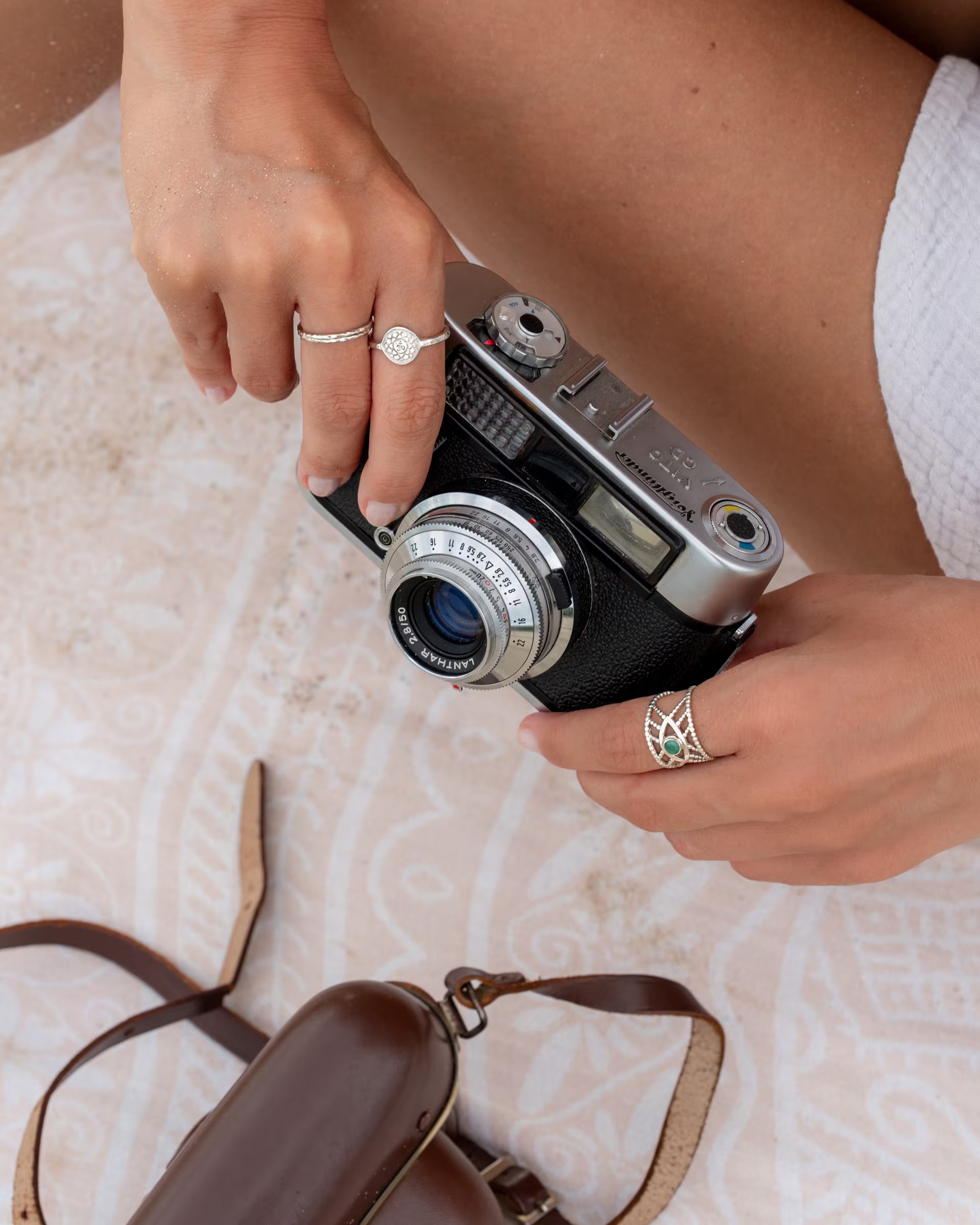 Nahaufnahme im Beach-Look von den Händen einer Frau, die eine Vintage-Kamera halten und mehrere auf Bali handgefertigte silberne Ringe – darunter der breite Intuition Ring mit grünem Smaragd-Edelstein sowie ein offener MOONPHASES CHANDRA-Ring – aus 925er Silber von der Schmuckmarke ILUH tragen.