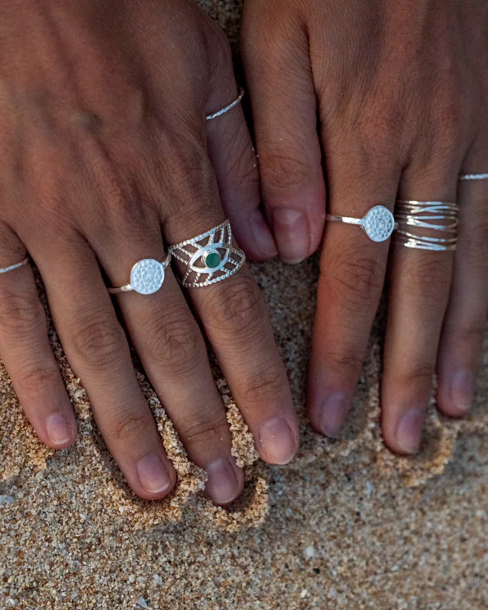 Handgefertigtes Silberschmuck-Set aus Bali von der Marke ILUH, bestehend aus CHAKRA-Motivringen, dem INTUITION-Ring mit grünem Smaragd-Edelstein, dem SARI-Ring und dem fein gehämmerten AURA-Ring aus 925er Silber; präsentiert an den Händen einer Frau im Sand am Strand, eingefangen nach dem Sonnenuntergang bei stimmungsvollem Abendlicht auf Bali.