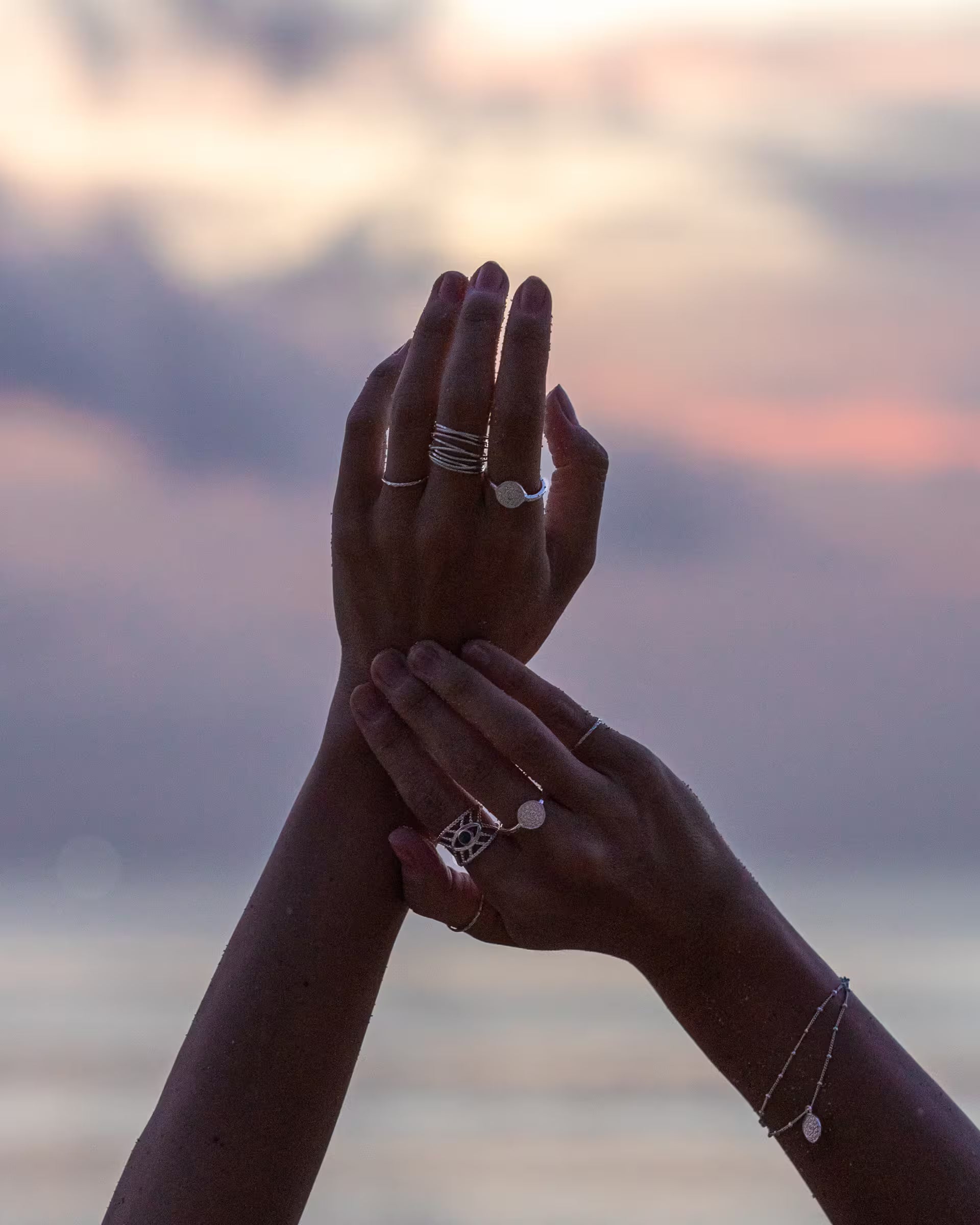 Stimmungsvolle Nahaufnahme im Beach-Look von zwei elegant emporgehobenen Händen vor einem malerischen Sonnenuntergang am Meer, die eine Kombination aus mehreren auf Bali handgefertigten silbernen Chakra Ringen und ein feines Chakra Armband aus 925er Silber von der Schmuckmarke ILUH tragen.