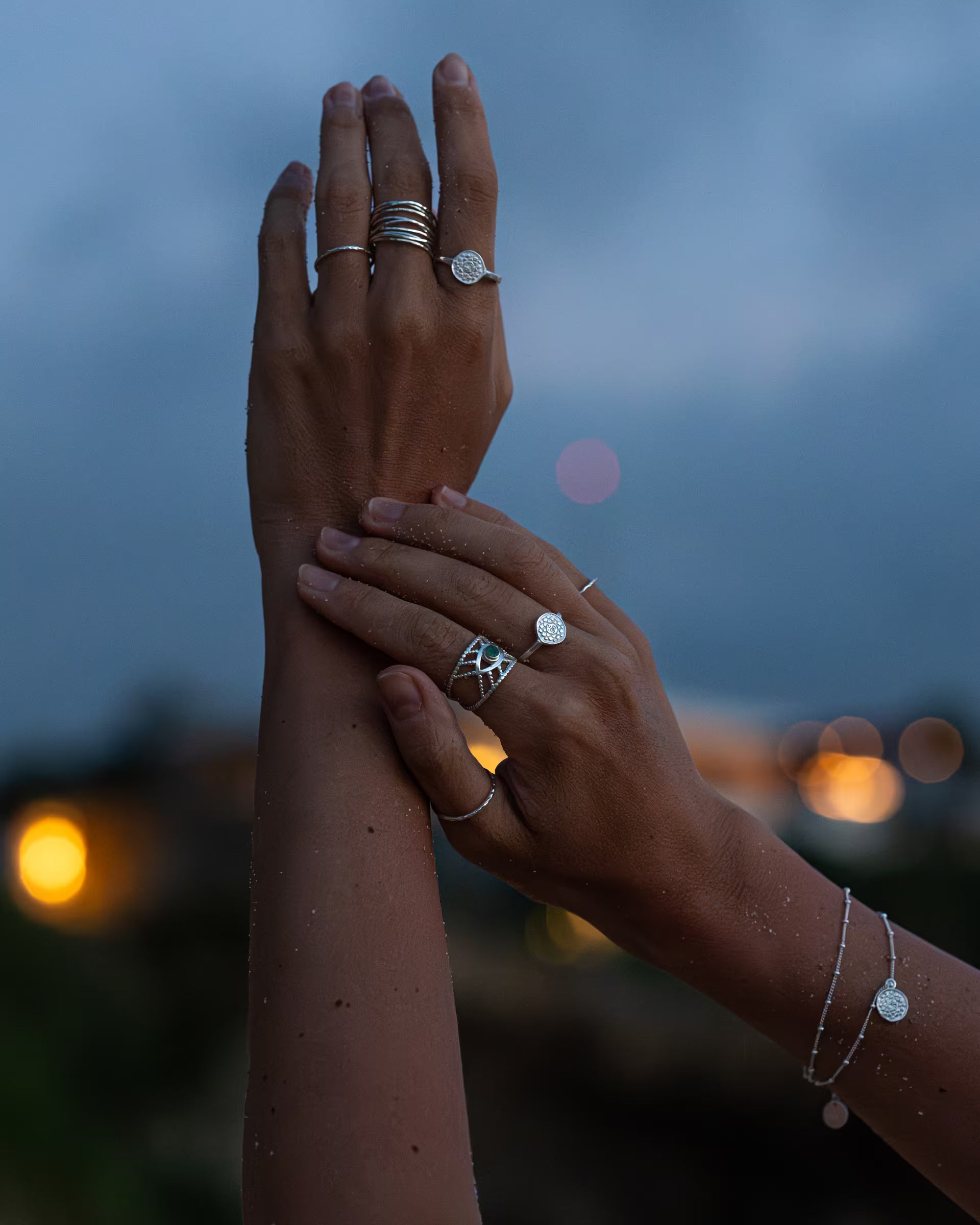 Auf Bali handgefertigte silberne Ringe, darunter der Intuition Ring mit grünem Smaragd-Edelstein, und das passende Chakra Armband und Chakra Ringe aus 925er Silber von der Schmuckmarke ILUH, getragen an den Händen einer jungen Frau bei Dämmerung mit weichem Lichter-Bokeh im Hintergrund auf Bali.