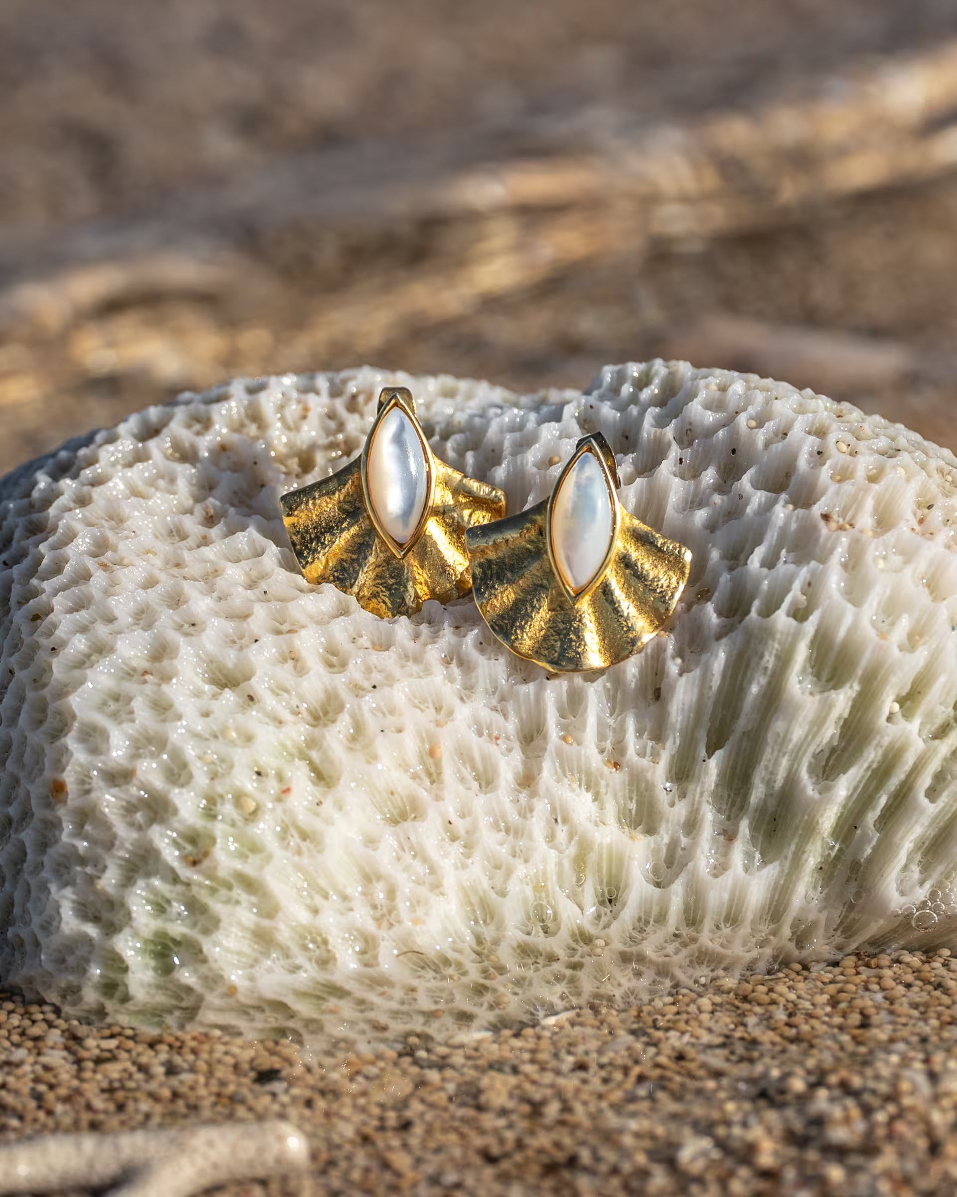 Handgefertigte, 18k vergoldete ALUN Stecker-Ohrringe aus 925er Silber in Muschel-Optik mit schimmerndem Perlmutt von der Schmuckmarke ILUH, stilvoll platziert auf einer hellen Koralle im Sand am Strand.