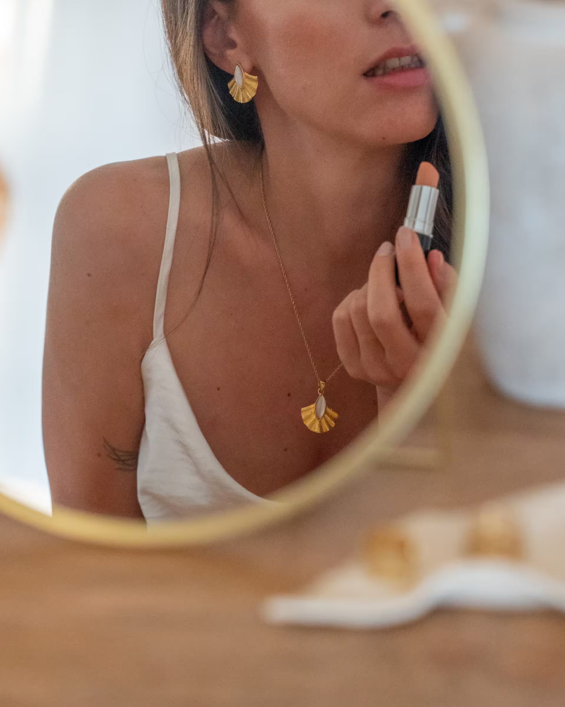 Junge Frau betrachet sich beim Auftragen von Lippenstift im Spiegel und trägt dabei goldene auf Bali handgefertigte Alun Kette und Alun Ohrringe von ILUH