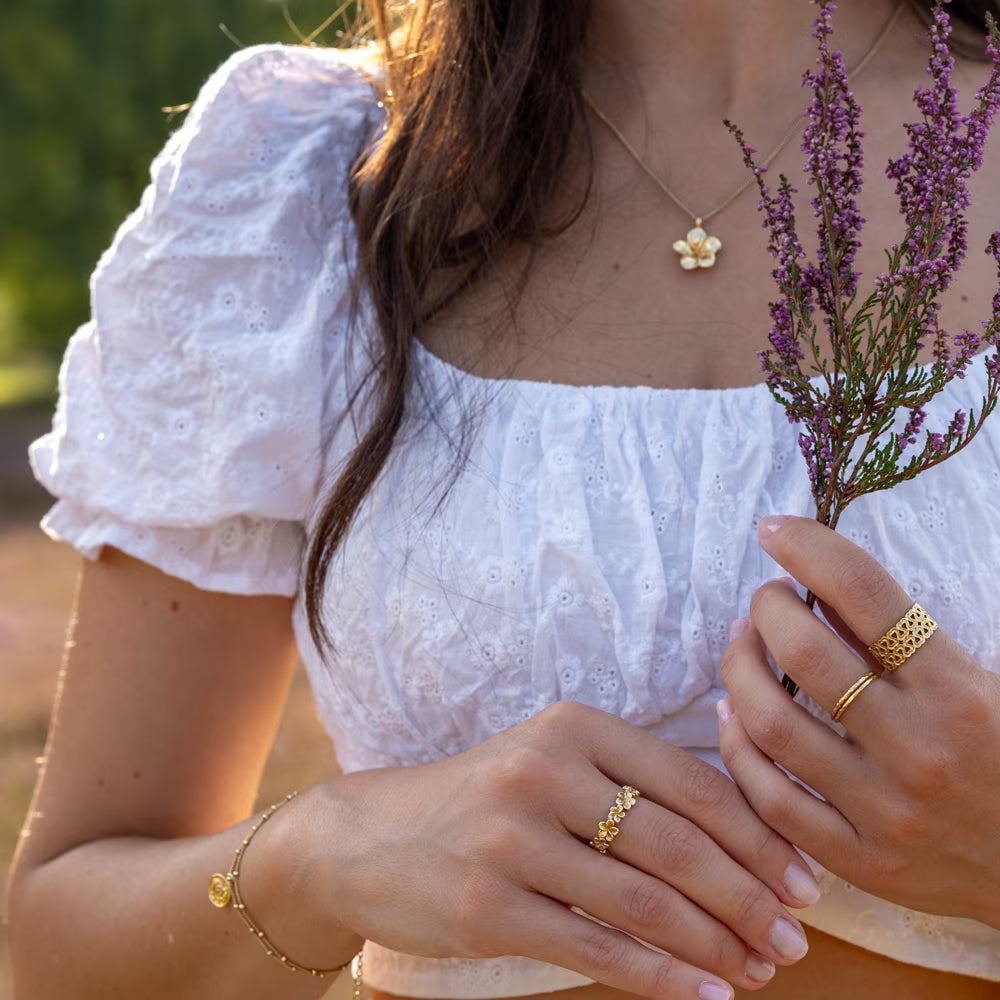 Ein romantischer, verspielter Spätsommer-Look: Eine junge Frau in weißem Broderie-Anglaise-Top hält zartes Heidekraut. Ihr Look wird durch die filigranen ILUH Jewelry Ringe, das Armband und die Halskette aus 925 Sterling Silber mit 18k Vergoldung vollendet – fair und nachhaltig in Bali handgefertigt.